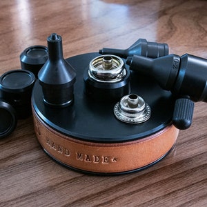 Könnte beinhalten: Eine schwarze, lederbezogene Basis mit der Gravur "HAND MADE*" . Die Basis verfügt über eine Vielzahl von Werkzeugen zum Setzen von Ledernieten, darunter einen schwarzen Metallstanz, ein schwarzes Metallsetzwerkzeug und einen silbernen Metallniet.