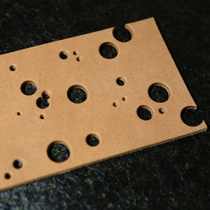May include: A brown leather sheet with multiple circular cutouts of varying sizes. The sheet has a rough texture and is lying on a black surface.