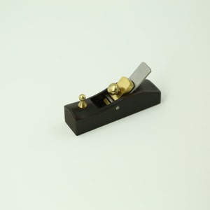 Op de afbeelding: Een kleine, zwarte houten handvlak met een messingknop en een metalen mes. De vlak is ontworpen voor houtbewerking en is een handig hulpmiddel voor het gladstrijken en vormen van hout.