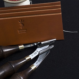 Puede incluir: Tres herramientas de trabajo de cuero con mangos de madera y cuchillas de metal. Las herramientas están sobre una superficie de tela negra. Un trozo de cuero marrón con el texto "YORKSHIRE hand made" está en el fondo.