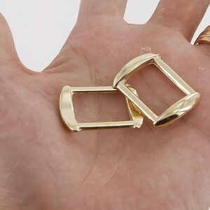Puede incluir: Dos hebillas rectangulares doradas con bordes redondeados. Las hebillas son de metal brillante y se muestran sobre un fondo claro. Las hebillas son probablemente para usar en cinturones, bolsos u otros accesorios.