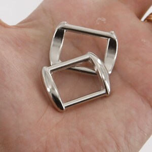 Puede incluir: Dos hebillas met&aacute;licas plateadas y brillantes. Cada hebilla tiene forma rectangular con bordes redondeados y una barra en el centro. Las hebillas se muestran sobre un fondo claro.