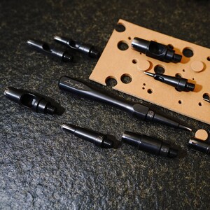 May include: A set of leather hole punch tools, including a long handled punch with the text "Dury Leather" on the handle, and several smaller punches. The tools are on a dark gray textured surface with a brown leather cutting mat in the background.