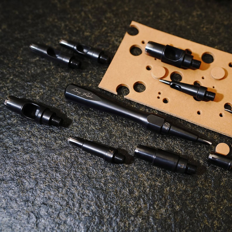 May include: A set of leather hole punch tools, including a long handled punch with the text "Dury Leather" on the handle, and several smaller punches. The tools are on a dark gray textured surface with a brown leather cutting mat in the background.
