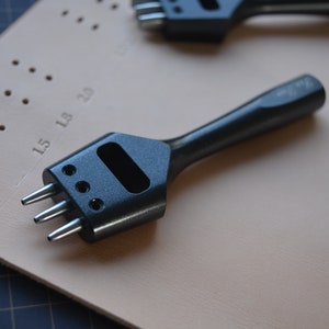 May include: A black leather punch tool with a handle and three prongs. The tool is lying on a light brown leather surface with a grid pattern.
