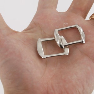 May include: Two silver metal D-rings, also known as D rings or D-shaped rings, are shown in a hand. The rings are stacked on top of each other.