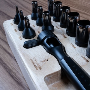 Op de afbeelding: Een set zwarte leren ponsen in een houten houder. De ponsen zijn in een rij gerangschikt en zijn gelabeld met hun maten in millimeters. De houder heeft de tekst "YUKESHINE HANDY BARK" gegraveerd.
