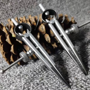 Könnte beinhalten: Zwei silberne Zeichenkompasse aus Metall mit verstellbaren Spitzen. Die Kompasse liegen auf einer braunen Tannenzapfen.