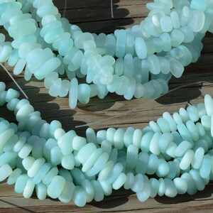 May include: A strand of light blue, opaque, smooth, pebble-shaped beads. The beads are strung on a clear thread and are arranged in a loose, flowing pattern.