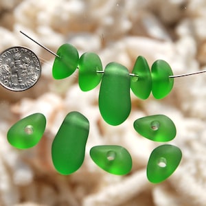 May include: A collection of seven green sea glass beads with holes drilled through the center. The beads are various shapes and sizes, and some are strung on a wire.