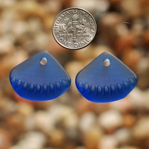 May include: Two blue glass sea glass pendants with a scalloped edge and a hole for stringing. The pendants are about the size of a dime.