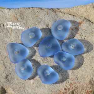 May include: Ten light blue glass beads with a hole in the center. The beads are irregularly shaped and have a frosted finish. The beads are arranged on a light brown rock surface.