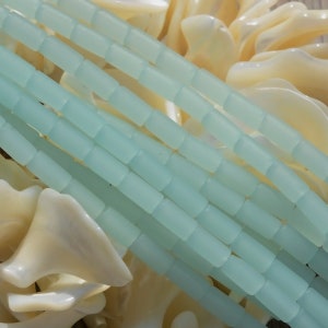 May include: A strand of light blue glass beads, each bead is a rectangular shape. The beads are arranged in rows and are stacked on top of each other. The beads are on a white background.