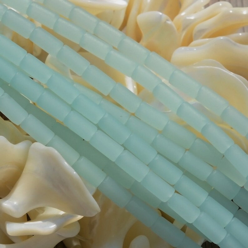 May include: A strand of light blue glass beads, each bead is a rectangular shape. The beads are arranged in rows and are stacked on top of each other. The beads are on a white background.