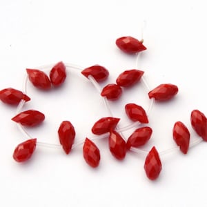 May include: A strand of red gemstone beads, each bead is faceted and shaped like a teardrop. The beads are strung on a clear elastic cord.