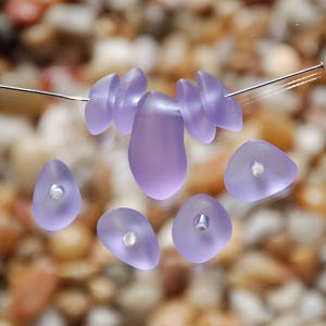 May include: A set of seven purple glass beads with a hole in the center of each bead. The beads are various shapes, including teardrop, oval, and triangular.