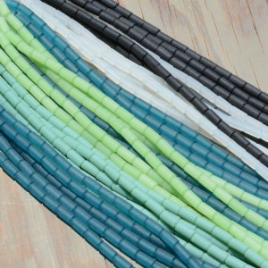 May include: A close-up of a variety of glass beads in shades of green and blue. The beads are small and cylindrical, and they are arranged in a random pattern. The beads are perfect for jewelry making or other craft projects.