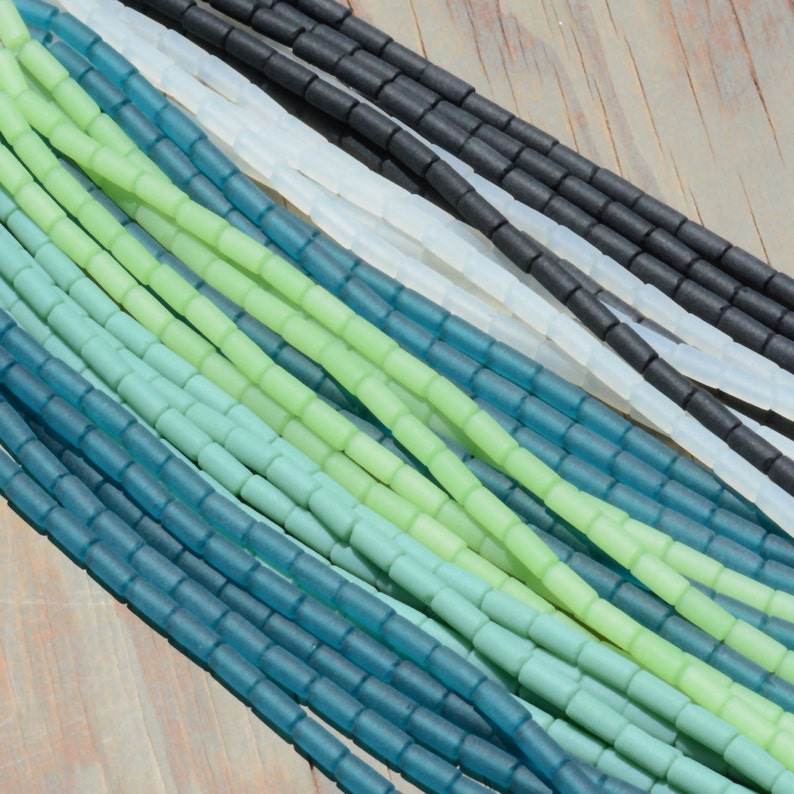 May include: A close-up of a variety of glass beads in shades of green and blue. The beads are small and cylindrical, and they are arranged in a random pattern. The beads are perfect for jewelry making or other craft projects.