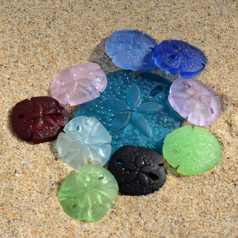 Pu&ograve; includere: Assortimento di conchiglie di vetro colorate in varie forme e dimensioni. Le conchiglie sono disposte su una superficie sabbiosa.