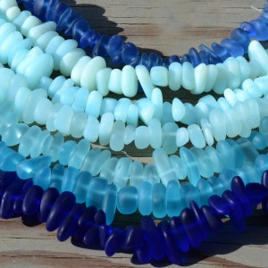 May include: A strand of blue and turquoise sea glass beads, with a darker blue strand at the bottom. The beads are all different shapes and sizes, and they have a frosted, translucent finish.