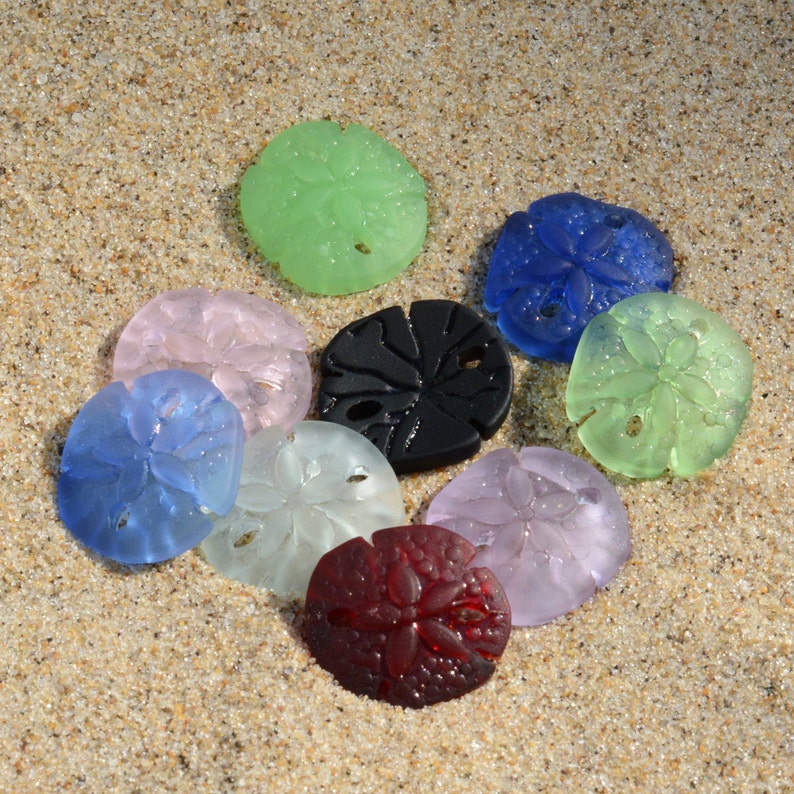 Pu&ograve; includere: Assortimento di nove ornamenti di vetro a forma di dollaro di sabbia in vari colori, tra cui verde, blu, rosa, bianco, nero e rosso. Gli ornamenti sono disposti su una superficie sabbiosa.