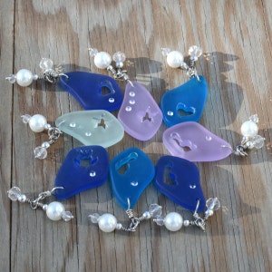 May include: A set of eight sea glass charms with various shapes and designs, including a heart, a butterfly, and a fish. Each charm is strung on a silver chain with a white pearl and crystal beads.