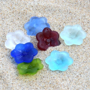 Pu&ograve; includere: Sette ornamenti di vetro a forma di fiore in vari colori, tra cui blu, bianco, rosso, verde e azzurro. Gli ornamenti sono disposti su una superficie sabbiosa.