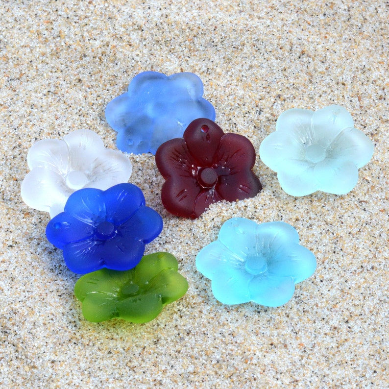 Pu&ograve; includere: Sette ornamenti di vetro a forma di fiore in vari colori, tra cui blu, bianco, rosso, verde e azzurro. Gli ornamenti sono disposti su una superficie sabbiosa.