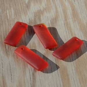 Op de afbeelding: Vier rechthoekige hangers van rood glas met een gat aan de bovenkant. De hangers liggen op een houten oppervlak.