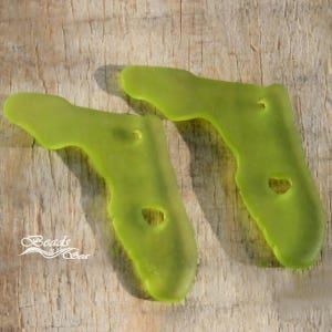 May include: Two pieces of sea glass in a bright green color. The glass is smooth and has been tumbled by the ocean. The glass is shaped like a foot.