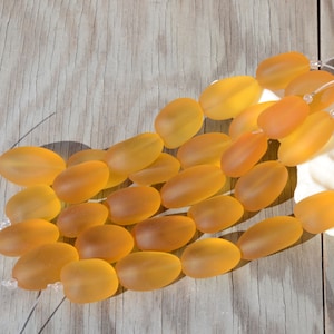 May include: A collection of smooth, frosted glass beads in a warm amber color. The beads are oval shaped and have a slightly translucent appearance.