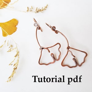 May include: A pair of copper wire earrings shaped like ginkgo leaves. The earrings are hanging from a white background with dried leaves and grass. The text "Tutorial pdf" is written below the earrings.