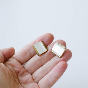 Könnte beinhalten: Ein Paar quadratische Ohrringe mit goldfarbenem Rahmen und weißer Mitte. Die Ohrringe werden in einer Hand vor weißem Hintergrund gehalten. Die Ohrringe haben ein schlichtes, elegantes Design.