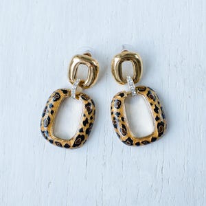 Puede incluir: Un par de pendientes dorados con un diseño rectangular de estampado de leopardo. Los pendientes tienen un lazo dorado en la parte superior y un lazo más pequeño con cristales transparentes que conecta con el rectángulo con estampado de leopardo.