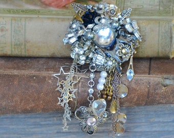 May include: A silver and white vintage brooch with a dangling chain of stars and beads. The brooch is made of various materials, including metal, glass, and pearls. It is sitting on top of two old books.