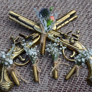 May include: Five gold-toned, decorative, miniature pistols with small bouquets of white flowers attached to each. The pistols are crossed in the center of the image, with the bouquets arranged around them. The pistols are on a brown, woven background.