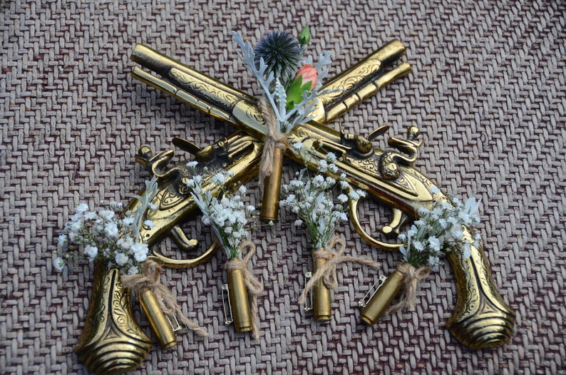 May include: Five gold-toned, decorative, miniature pistols with small bouquets of white flowers attached to each. The pistols are crossed in the center of the image, with the bouquets arranged around them. The pistols are on a brown, woven background.