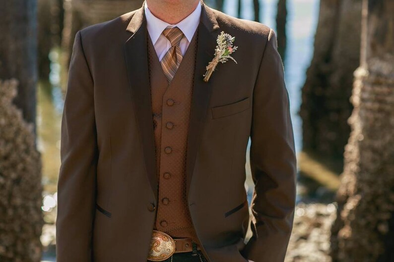 May include: A brown suit jacket with a brown vest and a brown and white patterned tie. The jacket has a single button closure and a pocket on the left side. The vest has a button closure and a pocket on the right side. The tie has a floral boutonniere on the left side.