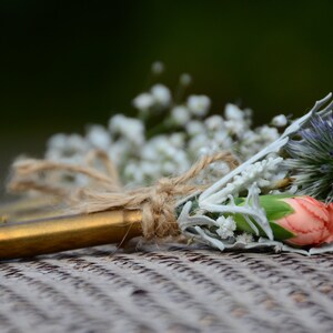 May include: A rustic wedding boutonniere made with a bullet casing, baby's breath, a single pink carnation, and a blue thistle.