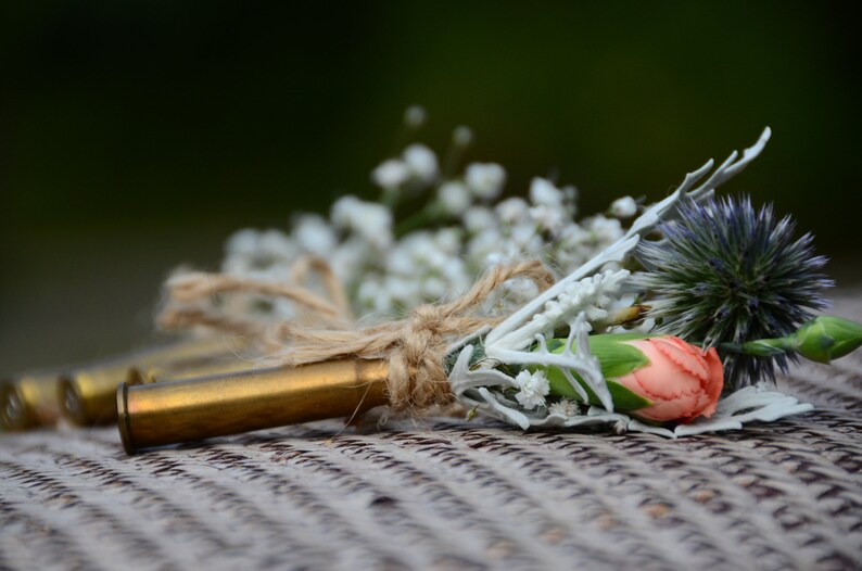 May include: A rustic wedding boutonniere made with a bullet casing, baby's breath, a single pink carnation, and a blue thistle.