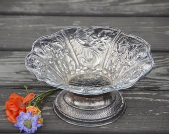 May include: A silver bowl with an ornate design and a pedestal base. The bowl is on a wooden surface with flowers next to it.