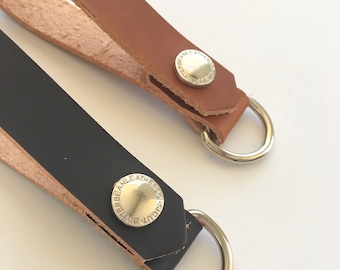 May include: Two leather keychains with silver metal rings and a silver button with the text "BUTTERBEAN LEATHERCO" on each. The keychain on the left is black leather and the keychain on the right is brown leather.