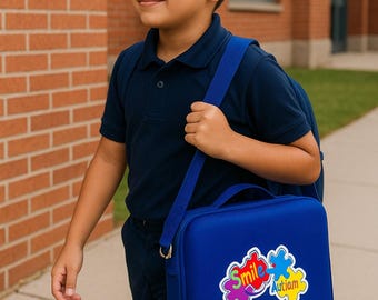 May include: A young person wearing a navy blue polo shirt and pants, carrying a blue lunch bag with a shoulder strap. The bag has a colorful puzzle piece design with the words "Smile" and "Autism". The background includes a brick building.