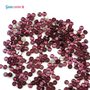 May include: A large quantity of small, round, red-purple glass beads. The beads are scattered on a white surface.