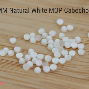 May include: A close-up of a group of 2mm white mother of pearl cabochons on a wooden surface. The cabochons are round and have a smooth, polished finish.