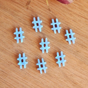 May include: Eight blue opal hashtag charms with a sparkly finish. The charms are arranged in a circle on a wooden surface.