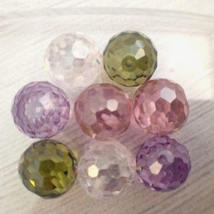 May include: A collection of eight faceted glass beads in various shades of purple, pink, and green. The beads are round and have a shiny, iridescent finish.