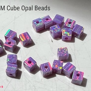 May include: A collection of 4mm cube opal beads. The beads are a light purple color with iridescent flashes of color. Each cube has a hole drilled through the center. The text "4MM Cube Opal Beads" is at the top.