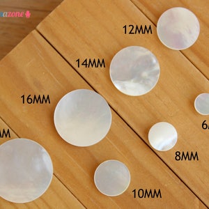 May include: Assortment of white mother of pearl round buttons in various sizes, ranging from 6mm to 18mm. The buttons are arranged on a wooden surface.