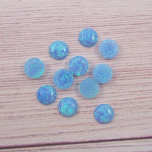 May include: A collection of 10 round, light blue opal cabochons. The cabochons are scattered on a light brown wooden surface.